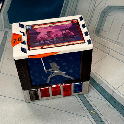 May include: A multi-colored game piece with a card featuring a landscape scene and the text "The Nest". The piece has a blue and white top, a black and silver body, and an orange dial with the number 70. A blue panel displays a silhouetted figure.